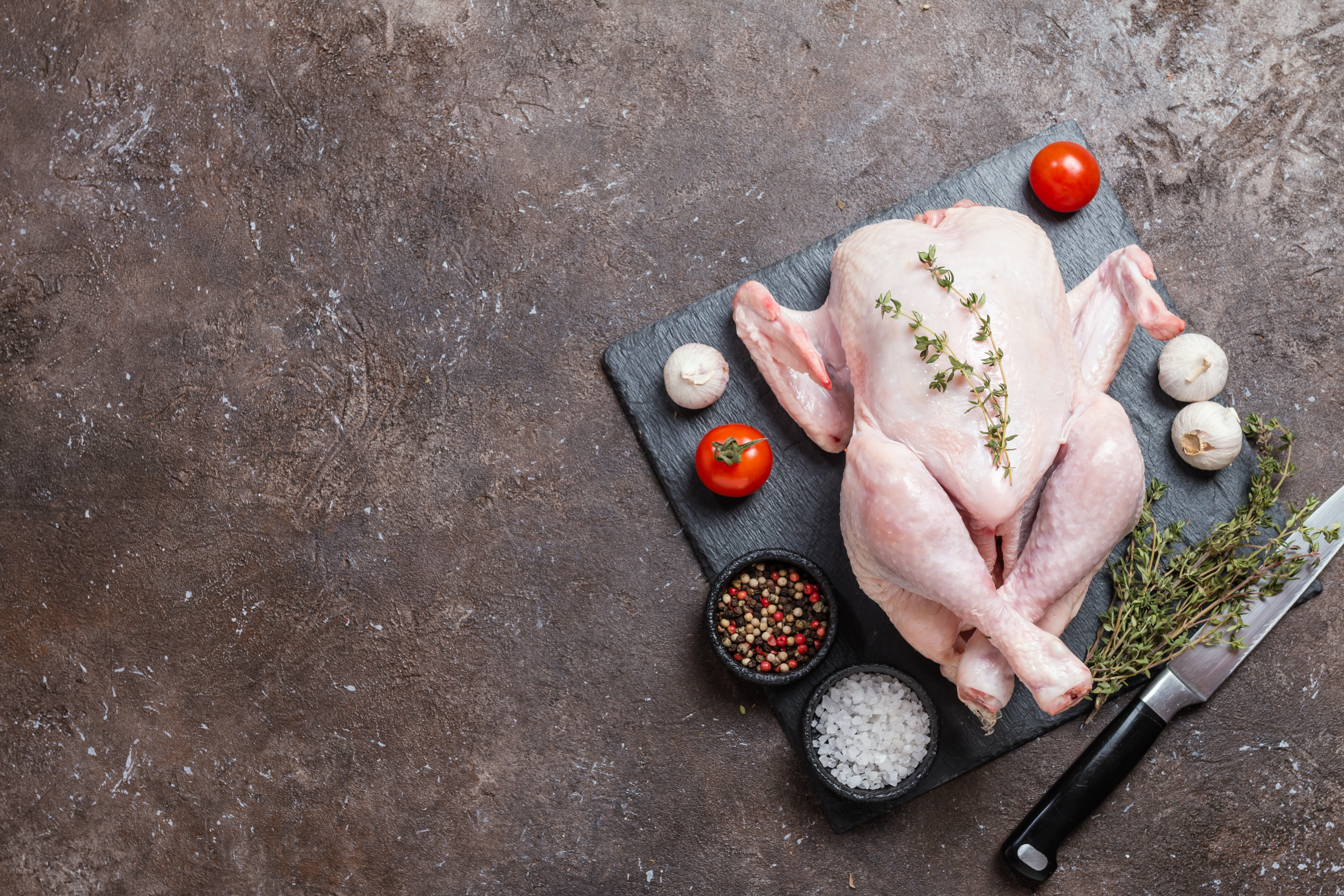 Raw whole chicken on a slate cutting board, surrounded by fresh herbs, tomatoes, garlic, and bowls of mixed peppercorns and sea salt, with a knife nearby, ideal for culinary preparation.