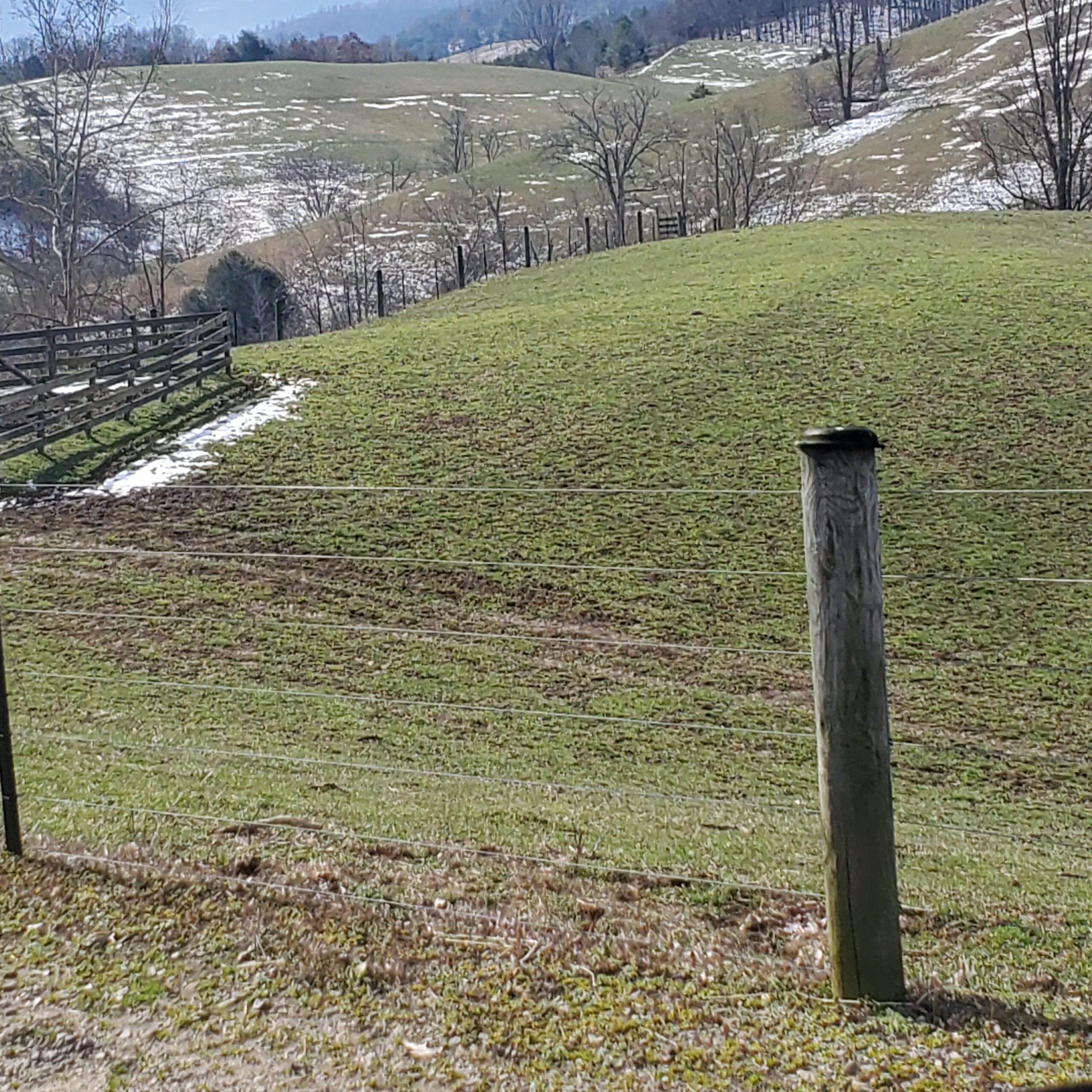 Scenic view of rolling green hills with patches of snow, a wooden fence along the edge, and a wooden fence post in the foreground, showcasing a tranquil rural landscape.